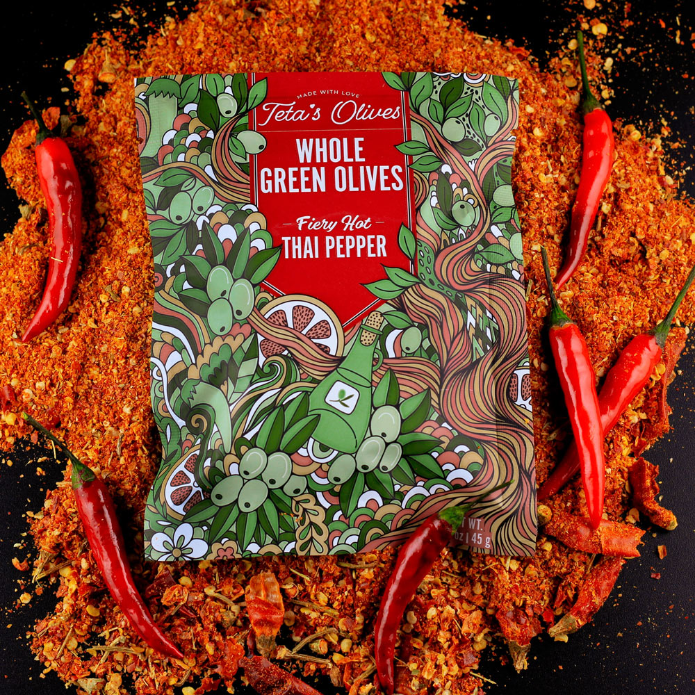 image of a bag of teta's olives fiery hot thai pepper laying on a bed of crushed red peppers, surrounded by red thai peppers
