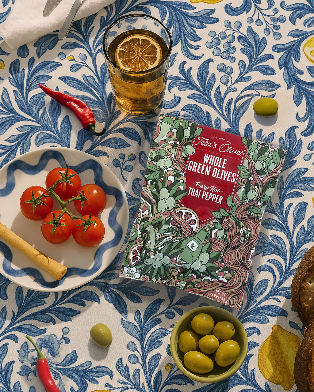 Overhead picture of Teta's Olives Thai Pepper on a table surrounded by green olives in a bowl, tomatoes, peppers, a lemonade cup