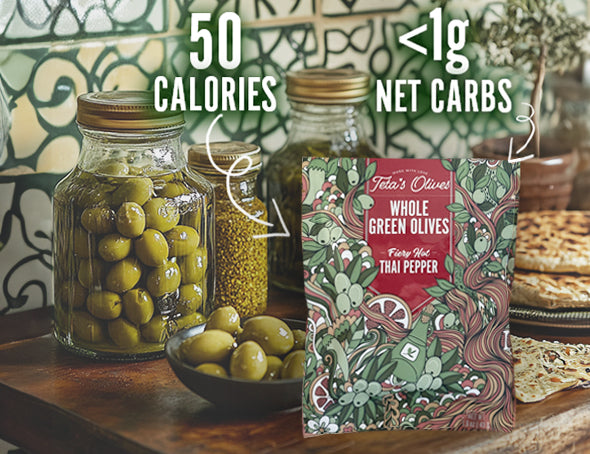 front view of teta's olives fiery hot thai pepper snack bag on a kitchen counter with jars and a bowl of green olives. text saying "50 calories" and "<1g net carbs" pointing at the bag