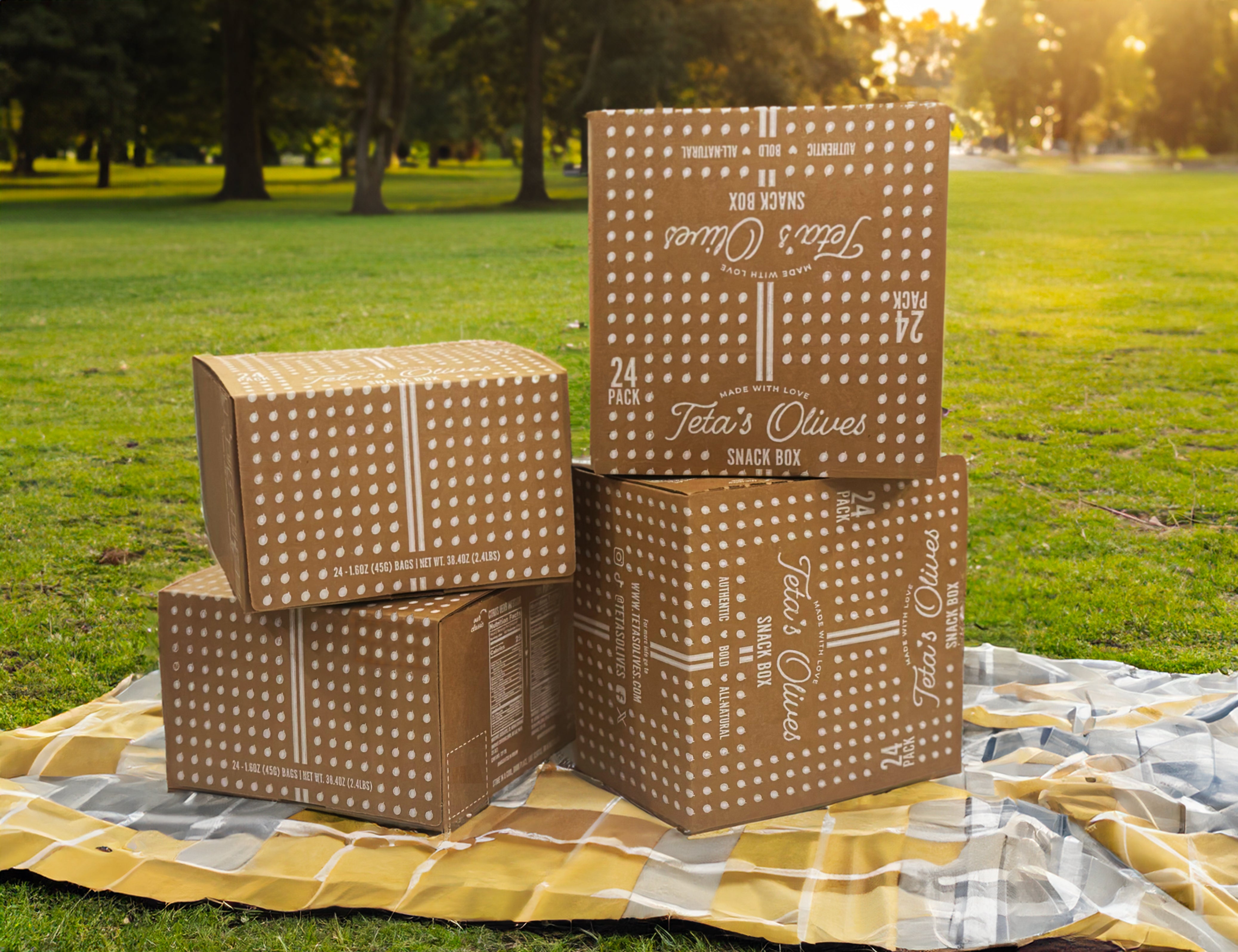 stacked boxes of teta's olives 24-pack snack boxes on a picnic blanket in the park